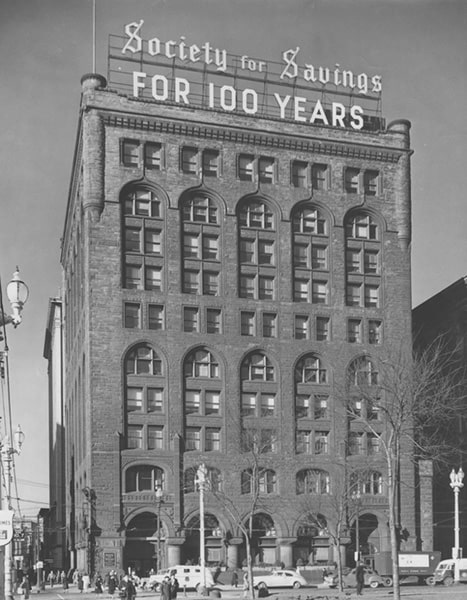 A black-and-white photograph of a ten-story building with a large sign on top of the building stating 'Society for Savings for 100 years.'